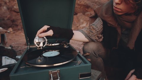 Tilt up shot of young woman in worn clothes starting vinyl record player, enjoying music and dancing at campsite in post apocalyptic world