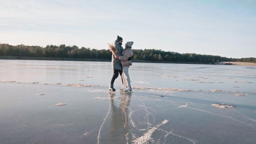 Cute couple kisses standing on a frozen lake on a sunny winter day with beautiful landscape. Man and woman having a date at the winter lake. Ice skating on the lake. High quality 4k footage