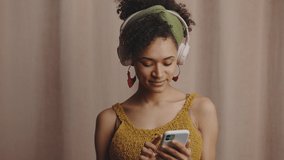 Young happy african american lady in headphones choosing music on mobile app and dancing, recording video for social media, beige studio background, slow motion