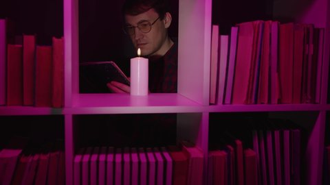 Serious man reading book near candle in bookcase. Male bookworm in eyeglasses reading book near burning candle placed on shelf of wooden bookcase with various literature in pink illumination