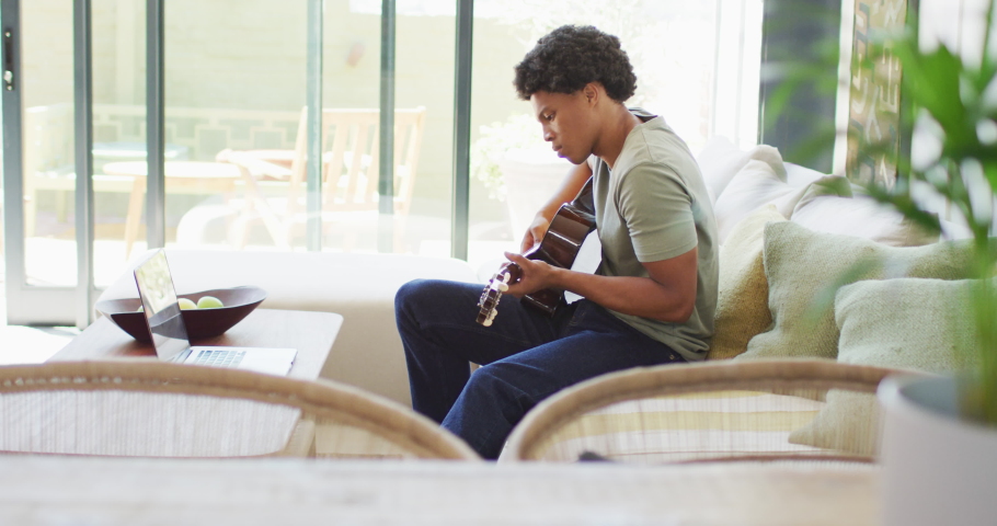 African american man plays guitar and singing, using laptop at home. leisure time using technology, relaxing at home alone.