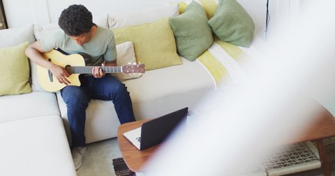 African american man plays guitar and singing, using laptop at home. leisure time using technology, relaxing at home alone.