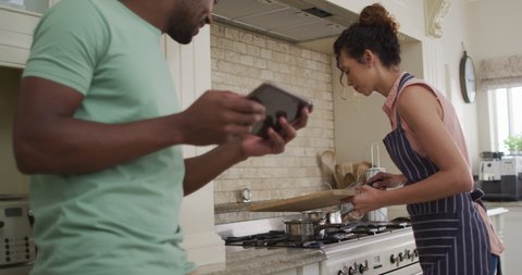 Happy biracial woman putting vegetables into saucepan and talking to her partner holding tablet. love and relationship, enjoying quality leisure time at home.