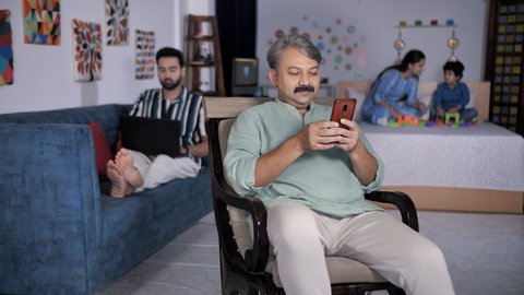 Indian family at home - three generations in one frame. Old man browsing his mobile phone. A beautiful mother playing with her young son while sitting on the bed - leisure time, parent-child bondin...