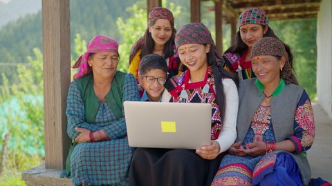A cheerful Asian indian group of family members including women wearing traditional costumes and a kid sitting together using a laptop in a remote or rural area. Concept of distant communication.