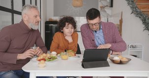 Happy gay family adoptive parents and curly-haired step son enjoy breakfast watching videos on tablet at kitchen table at home slow motion