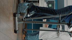 Vertical video: Elder person with mechanical disorders using stationary bicycle to treat musculoskeletal system, training to increase mobility. Female nurse assisting old patient at physical therapy.