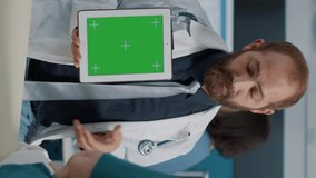 Vertical video: General practitioner using digital tablet with green screen at medical examination with patient. Doctor holding isolated mockup background with chroma key template and blank copy space