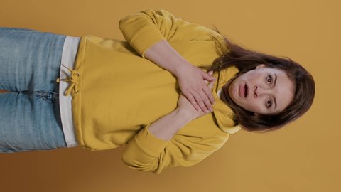 Woman having panic attack feeling short of breath getting scared and anxious in studio. Panicked casual young adult wearing hoodie choking because of shock and anxiety over yellow background.