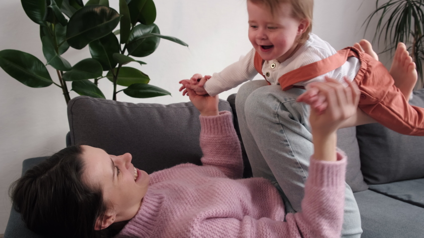 Caring young mom lying on comfortable sofa on back holding little baby daughter in air on legs bent in knees. Carefree child girl act flying doing gymnastics with mom. Play game imitate flight concept Royalty-Free Stock Footage #1086869606