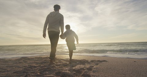 Back view of hispanic father and son walking on beach at sunset. family holidays, free time, togetherness and spending time outdoors.