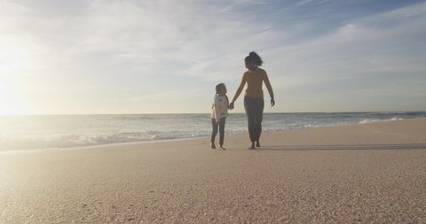 Happy hispanic mother and daughter walking on beach at sunset. family holidays, free time, togetherness and spending time outdoors.