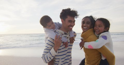 Portrait of happy hispanic parents carrying piggyback children on beach. family holidays, free time, togetherness and spending time outdoors.