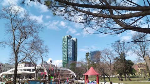 City park with playground in Tirana Albania, people enjoying sunny winter day. Kids playing and having fun on slide, swing and playhouse, a tower in the background, under blue sky. Feb 06.2022.