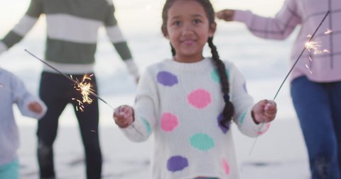 Portrait of happy hispanic girl playing with sparklers on beach with family at sunset. family holidays, free time and togetherness.