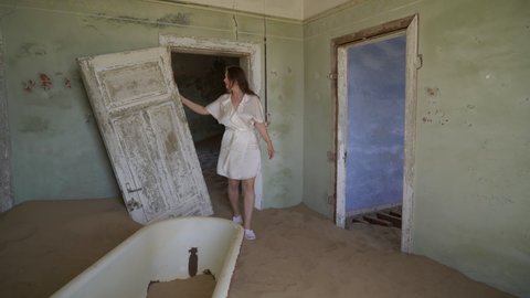 Young woman in robe in abandoned building in bathroom walks near bath tub. Ghost town Kolmanskop in desert. Room filled with sand. Old rusty bathtub in ruined house. Girl in white silk underwear.