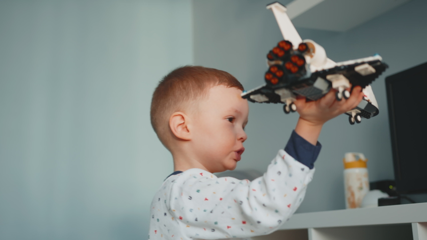 Portrait little 4 years caucasian blond boy in white pajamas is playing with space shuttle from constructor, holding it in his hands and flying in apartment. Boy flies with toy constructor spaceship. Royalty-Free Stock Footage #1086873734