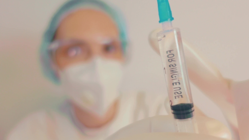 Extreme Close-up of a scientist, nurse or doctor in a white respirator, gloves and protective glasses, the developer of the coronavirus vaccine is holding a white transparent syringe. The doctor looks Royalty-Free Stock Footage #1086874310