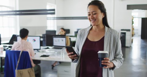 Happy asian businesswoman walking in office, holding takeaway coffee, using smartphone. independent creative business, working at a modern office.