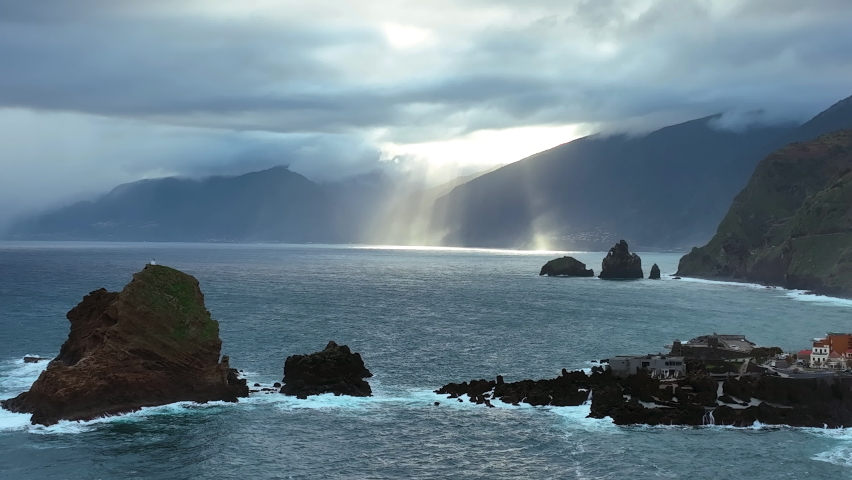 Beautiful sun beams go through the solid clouds. Porto Moniz town on Madeira island. Beautiful landscape with ocean bay covered with rocks. Small harbor on the rocks of Madeira.
