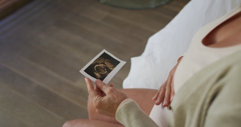 Hands of caucasian pregnant woman sitting on bed, touching belly and looking on ustrasound photo. pregnancy, expecting baby and spending time at home.