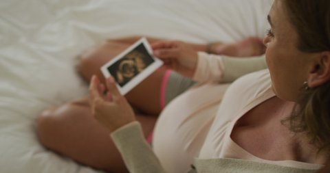 Caucasian pregnant woman sitting on bed, touching belly and looking on ultrasound photo. pregnancy, expecting baby and spending time at home.
