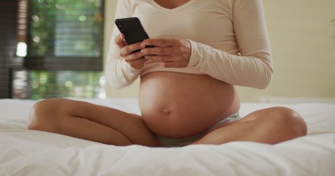 Midsection of caucasian pregnant woman sitting on bed and putting headphones on belly. pregnancy, motherhood, expecting baby, spending time at home and technology concept.