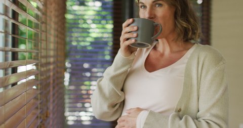 Happy caucasian pregnant woman standing at window with tea and touching belly. pregnancy, expecting baby and spending time at home.