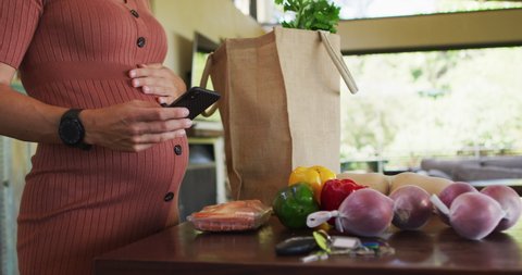 Midsection of happy caucasian pregnant woman touching belly, using smartphone in kitchen. expecting baby and healthy lifestyle during pregnancy.