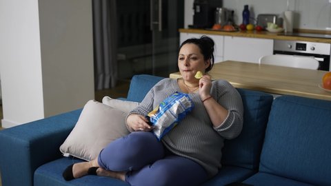 Overweight woman sitting on couch at home, watching tv and eating potato chips