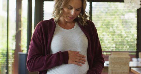 Portrait of happy caucasian pregnant woman touching belly and looking at camera. pregnancy, expecting baby and spending time at home.