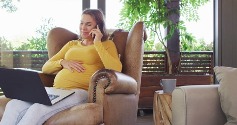 Happy caucasian pregnant woman sitting in armchair having phone call and using laptop. pregnancy, expecting baby, technology and spending time at home.