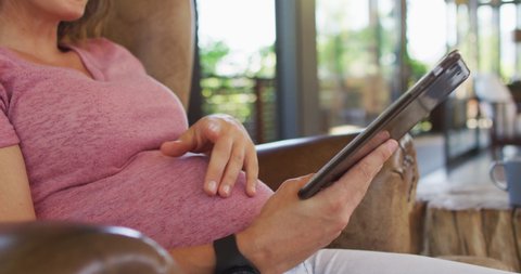 Midsection of caucasian pregnant woman sitting in armchair using tablet. pregnancy, motherhood, expecting baby, technology and spending time at home.