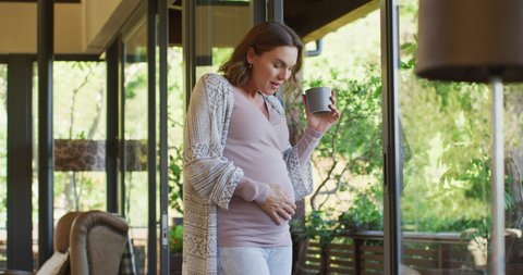 Happy caucasian pregnant woman standing at window with cup of tea and touching belly. pregnancy, motherhood, expecting baby and spending time at home.