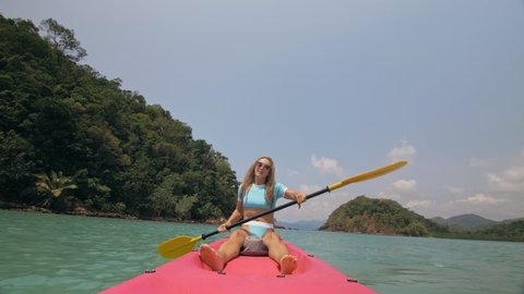 Pretty young blonde woman in stylish swimsuit and sunglasses sits on pink canoe sailing along sea against hills. Traveling to tropical countries. Positive sport girl hand padding on kayak front view.