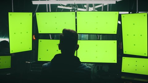Zoom out view of unrecognizable man using computers with chromakey green screens while creating virus in dark room of hacker base at night