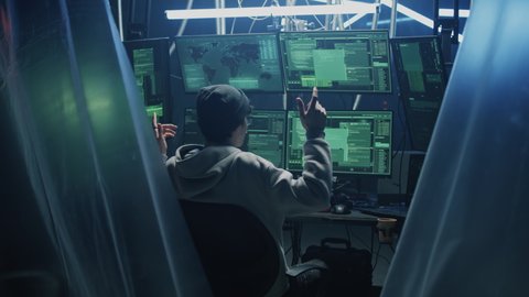 Zoom in view of happy young man in hoodie with glasses and hat dancing in excitement after successful hacker attack, while sitting at desk with computers in dark room