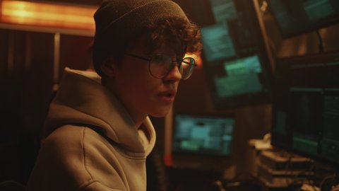 Young man hacker in hat and glasses talking with associate and using computers to hack data base while sitting at desk on dim criminal base