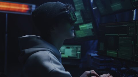 A teen hacker in a hat and augmented reality glasses siting at a desk in cybercriminal refuge near computers. He is trying to hack data or steal money in the meta universe