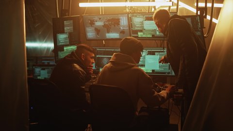 Zoom out view of young man in hoodie hacker helping associate to hack database for criminal while gathering around table in dim room