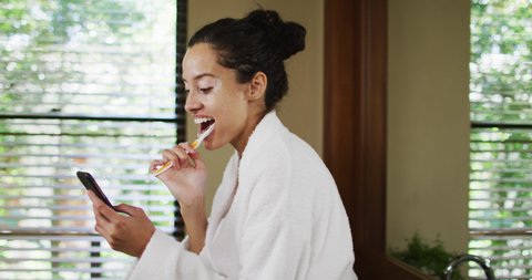Happy biracial woman in robe brushing teeth and using smartphone. beauty, wellness and self care, enjoying leisure time at home with technology.