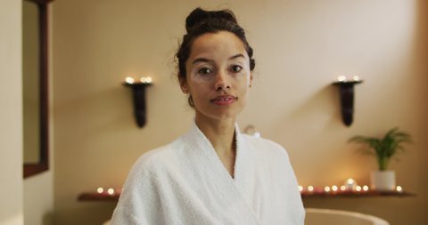 Portrait of happy biracial woman in robe standing in bathroom and looking at camera. beauty, wellness and self care, enjoying leisure time at home.