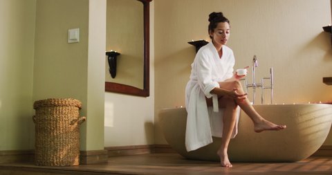 Happy biracial woman with vitiligo applying body lotion to legs in bathroom. beauty, wellness and self care, enjoying leisure time at home.