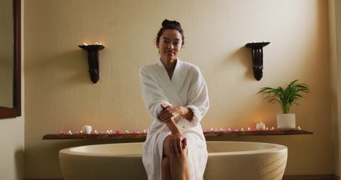 Happy biracial woman sitting in robe on bathtube. beauty, wellness and self care, enjoying leisure time at home.