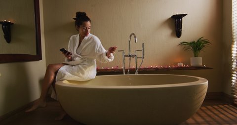 Happy biracial woman sitting in robe on bathtube, preparing bath, using smartphone. beauty, wellness and self care, enjoying leisure time at home with technology.