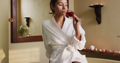 Relaxed biracial woman with vitiligo wearing bathrobe and drinking wine in bathroom. beauty, relaxation and self care, enjoying leisure time at home.