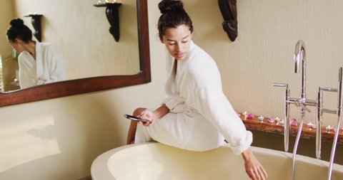 Happy biracial woman with vitiligo sitting in bathrobe, running bath and using smartphone. beauty, wellness and self care, enjoying leisure time at home with technology.