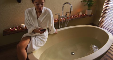 Happy biracial woman with vitiligo sitting in bathrobe, running bath and using smartphone. beauty, wellness and self care, enjoying leisure time at home with technology.