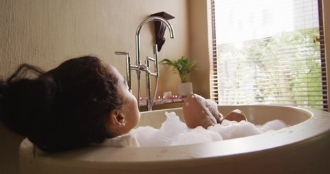 Happy biracial woman with vitiligo lying in bathtub, blowing foam and pampering herself. beauty, wellness and self care, enjoying leisure time at home.