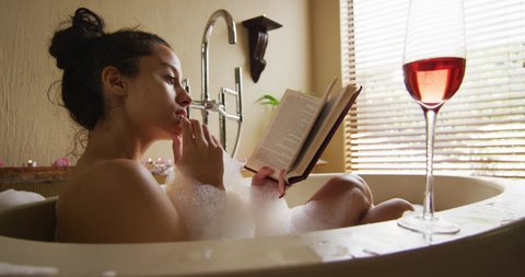 Relaxed biracial woman lying in bath with foam and glass of wine, reading book. beauty, wellness and self care, enjoying leisure time at home.